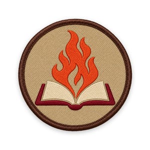 May include: A circular embroidered patch featuring a tan background with a dark brown stitched border. The design depicts an open book with cream-colored pages and a dark red cover, with a bright orange flame rising from the center of the book.