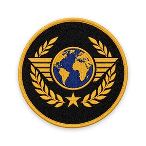 Puede incluir: Un parche bordado circular con fondo negro y borde dorado. El centro muestra un globo terráqueo azul con continentes dorados, flanqueado por alas doradas y enmarcado por una corona de laurel dorada con una estrella de cinco puntas abajo.
