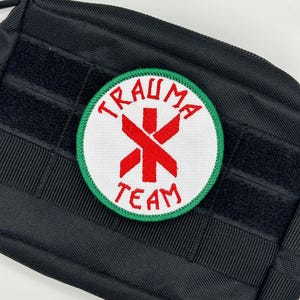 Könnte beinhalten: Ein runder, gestickter Aufnäher mit den Worten "TRAUMA TEAM" in roter Schrift auf weißem Grund, umrandet von einem grünen Rand. Der Aufnäher zeigt ein rotes Lebensstern-Symbol. Der Aufnäher ist an einer schwarzen taktischen Tasche befestigt.