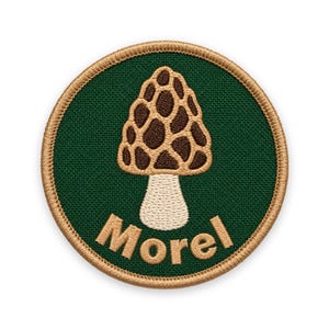 May include: A circular embroidered patch featuring a dark green background with a tan border. In the centre, a morel mushroom is depicted with a brown, honeycomb-textured cap and a cream-coloured stem. The word Morel is stitched in tan below.