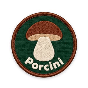 Puede incluir: Un parche bordado circular con un fondo verde oscuro y una ilustración de un hongo porcini de color marrón y crema en el centro. La palabra Porcini está bordada en letras mayúsculas blancas a lo largo del borde inferior, con un borde marrón.