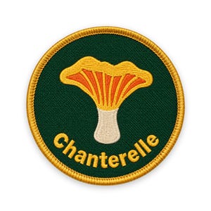 Peut inclure: Un écusson brodé circulaire avec un fond vert foncé et une illustration de champignon chanterelle jaune et orange au centre. Le mot Chanterelle est brodé en texte jaune sous le champignon, le tout fini par une bordure dorée.