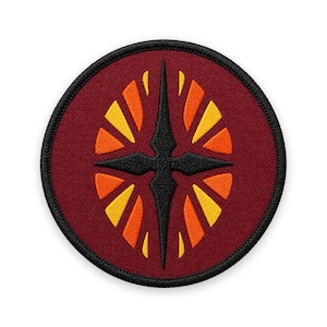 May include: A circular embroidered patch featuring a deep maroon background with a central black four-pointed star emblem. Surrounding the star are symmetrical, radiating segments in vibrant shades of yellow and orange, all enclosed by a thick black stitched border.