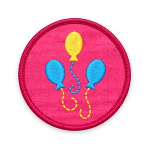 May include: A circular embroidered patch with a vibrant pink background. The design features one yellow balloon and two blue balloons, each with a matching stitched string. The patch has a thick, dark pink stitched border around the entire edge.