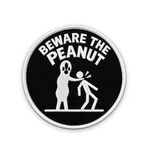 Peut inclure: Un écusson brodé circulaire avec un fond noir et une bordure blanche. Il présente un graphique blanc d'une grande créature en forme de cacahuète touchant le cou d'un bonhomme allumette. Le texte BEWARE THE PEANUT est arqué en blanc au-dessus.