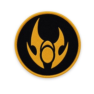 May include: A circular embroidered patch featuring a black background with a golden yellow border. In the center, a stylized golden yellow emblem depicts a central oval shape flanked by two curved, pointed forms that resemble flames or wings.