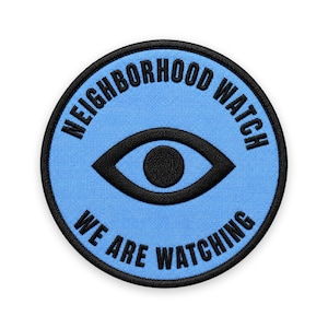 May include: A circular embroidered patch with a light blue background and a black border. The center features a black eye icon. The text "NEIGHBORHOOD WATCH" is arched across the top, and "WE ARE WATCHING" is arched across the bottom in black.