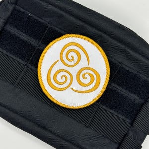 Può includere: Toppa rotonda con tre spirali dorate su sfondo bianco, bordata d'oro. La toppa è posta su uno sfondo nero strutturato, possibilmente una borsa o un'attrezzatura tattica.