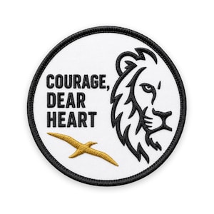 May include: A circular embroidered patch with a white background and a black stitched border. It features the black text COURAGE, DEAR HEART, a black lion profile illustration, and a small gold bird silhouette embroidered on the white fabric surface.
