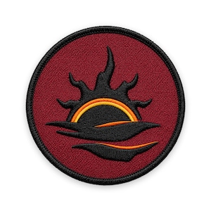 May include: A circular embroidered patch featuring a deep red background with a black border. The design depicts a stylized black sun rising over dark waves, accented by thin, curved orange and yellow lines that highlight the sun and water.