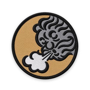 May include: A circular embroidered patch featuring a tan background with a black border. The design depicts a stylized gray face with wavy hair blowing a white cloud of air from its mouth, outlined in black thread against the tan fabric.