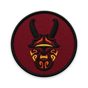 Può includere: Una toppa ricamata circolare con sfondo rosso scuro e bordo nero. Il centro presenta un design a maschera stilizzata in filo nero, giallo e arancione, caratterizzato da due corone prominenti e intricati motivi simmetrici.
