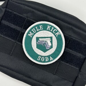 May include: A round embroidered patch with the text "MULE KICK SODA" in white lettering. The patch features a green border and a central design of a pistol. The patch is set against a black background.