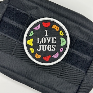 Peut inclure: Pochette tactique noire avec un écusson rond. L'écusson a une bordure blanche et porte l'inscription "I LOVE JUGS" en blanc, entourée de prises d'escalade colorées. La pochette est dotée d'une fermeture auto-agrippante.