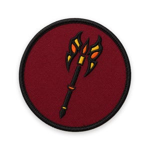 May include: A circular embroidered patch featuring a deep maroon background with a black border. Centered on the patch is a stylized black staff or weapon with orange and yellow flame-like accents at the top and along the handle shaft.