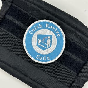 Pode incluir: Um patch redondo bordado com as palavras "Quick Revive Soda" em branco sobre um fundo azul claro. O patch apresenta um design de escudo com uma pessoa e uma lápide. O patch está em uma bolsa tática preta.