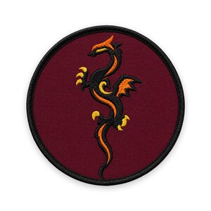 Puede incluir: Un parche bordado circular con un fondo granate oscuro y un borde negro. Centrado en el parche hay un diseño de dragón estilizado en negro, con acentos vibrantes en naranja y amarillo que resaltan sus alas, garras y cuerpo.