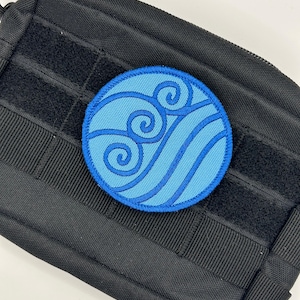Puede incluir: Un parche redondo bordado en azul con un símbolo de agua con líneas y olas arremolinadas. El parche está adherido a una bolsa táctica negra con correas de velcro y una cremallera.