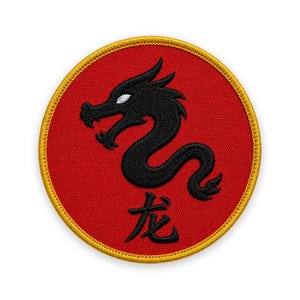 May include: A circular embroidered patch featuring a vibrant red background with a gold stitched border. Centered on the patch is a black dragon silhouette with a white eye, positioned above the black Chinese character for dragon, 龙.