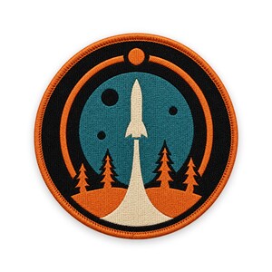 May include: A circular embroidered patch featuring a white rocket launching from an orange landscape with pine trees into a teal sky. The design includes black circular accents and a small orange planet, all bordered by a thick orange stitched edge.