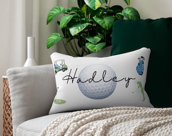 Könnte beinhalten: Weißes rechteckiges Kissen mit Golf-Motiv. Das Kissen zeigt eine Golfball-Grafik mit dem Namen "Hadley" in Schreibschrift sowie Illustrationen eines Golfwagens, einer Golftasche und eines Golfspielers. Das Kissen liegt auf einem grauen Sofa.