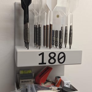 Może przedstawiać: Biały uchwyt na rzutki z różnymi rzutkami i akcesoriami. Uchwyt wyświetla numer "180" w dużych czarnych cyfrach. Akcesoria obejmują poziomicę, czerwone narzędzie Unicorn i zapasowe końcówki do rzutek.