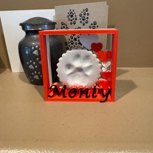 May include: A square orange frame containing a white circular paw print impression and the black text "Monty". The frame is decorated with several small red and white hearts. Behind the frame sits a dark gray urn with paw print patterns.