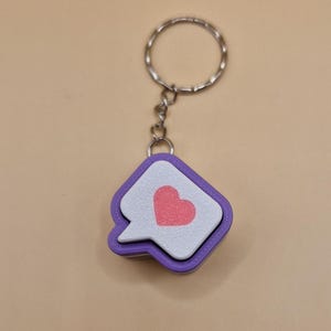 May include: A keychain with a purple, white, and pink heart design. The keychain has a silver ring and chain. The heart is inside a speech bubble shape.