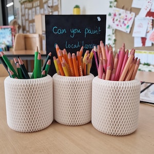 May include: Three cream-colored pencil holders with a diamond pattern, each filled with colored pencils. The pencils are green, orange, and pink. A chalkboard in the background reads "Can you paint local land".