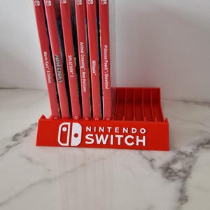 Op de afbeelding: Een rode Nintendo Switch game cartridge houder met het witte Nintendo Switch logo. De houder bevat verschillende game cases, waaronder "Mario Kart 8 Deluxe", "Minecraft" en "Splatoon 2".