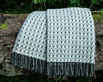 Puede incluir: Una manta plegada con un patrón de chevrón en tonos grises y blancos, con flecos en el borde inferior. La manta está colocada sobre un tronco en un entorno natural al aire libre, rodeada de vegetación verde.