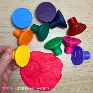 May include: A selection of brightly coloured emoji-shaped stampers in shades of blue, orange, yellow, green, red, pink, and purple. A hand holds a yellow stamper over red playdough, which has impressions of emoji faces. The stampers are for crafting.