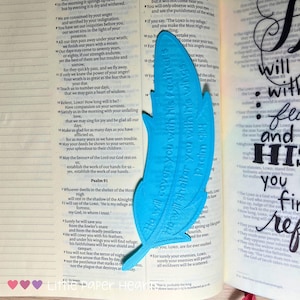 May include: A turquoise feather-shaped bookmark with the text "He will cover you with his feathers, He will shelter you with his wings, Psalm 91" rests on an open book. The book pages display printed text.