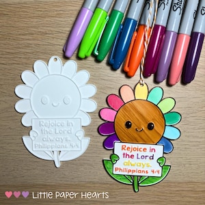 May include: Two sunflower-shaped ornaments, one white and one colourful, with the text "Rejoice in the Lord always. Philippians 4:4." A collection of colourful Sharpie markers are arranged above the ornaments. The ornaments are likely for crafts or gifts.