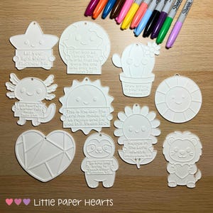 May include: A selection of white, unpainted, laser-cut shapes, including a star, globe, cactus, axolotl, sun, sunflower, heart, sloth, and lion. Each shape has a small hole for hanging and features inspirational Bible verses. Colourful markers are in the background.