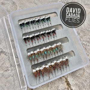 May include: A clear plastic case containing multiple fishing flies. The flies feature silver heads and colourful bodies, arranged in rows. The case rests on a textured stone surface. The text "DAVID CABADA FLIES" is in a black circle.