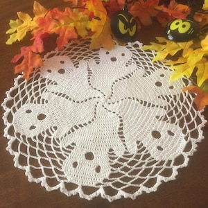 Peut inclure: Nappe blanche au crochet avec un motif de fantômes d'Halloween. La nappe présente six formes de fantômes avec des découpes pour les yeux et la bouche, disposées en cercle. La nappe est entourée de feuilles d'automne et de petites décorations de citrouilles noires.