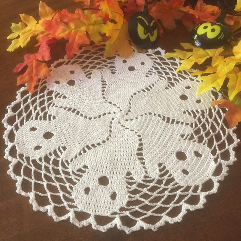 May include: A white, crocheted doily with a Halloween ghost design. The doily features six ghost shapes with eye and mouth cutouts, arranged in a circular pattern. The doily is surrounded by fall leaves and small black pumpkin decorations.