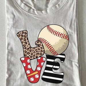 May include: A light beige t-shirt featuring a graphic design that spells the word LOVE. The letter L has a brown leopard print, the letter O is a baseball, the letter V is red with white baseball patterns, and the letter E has black and white stripes.