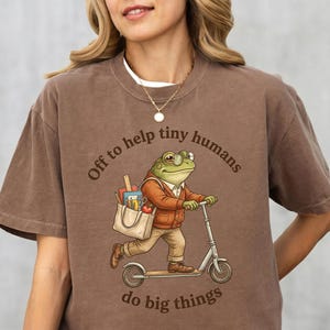 Puede incluir: Una camiseta marrón de manga corta con el gráfico de una rana con gafas y cárdigan, montada en un patinete y llevando una bolsa con material escolar. El texto dice, Off to help tiny humans do big things.