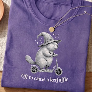 Könnte beinhalten: Ein violettes T-Shirt mit einer schwarz-weißen Grafik eines Bibers, der einen mit Sternen verzierten Zaubererhut trägt und auf einem Tretroller fährt. Unter der Illustration steht der Text "Off to cause a kerfuffle" in weißer Schrift.