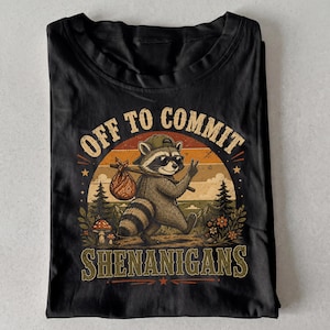 May include: A black t-shirt featuring a graphic of a raccoon wearing sunglasses and a cap, carrying a bindle on a stick. The design includes a sunset background, pine trees, mushrooms, and the text "OFF TO COMMIT SHENANIGANS" in a vintage font.