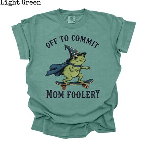 May include: A light green t-shirt featuring a graphic of a green frog wearing a blue wizard hat with yellow stars and a blue cape, riding a skateboard. The text "OFF TO COMMIT" is printed above the frog and "MOM FOOLERY" is printed below it.