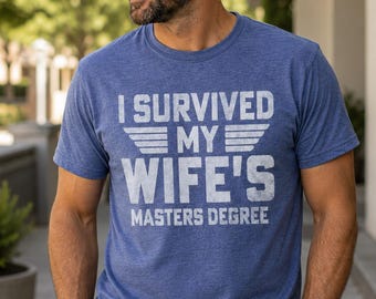 May include: A heather blue short-sleeve t-shirt featuring white distressed text that reads I SURVIVED MY WIFE'S MASTERS DEGREE with horizontal wing graphics flanking the word MY. The shirt has a crew neckline and a standard fit.