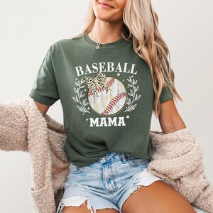 May include: An olive green short-sleeve t-shirt featuring a graphic of a baseball with a leopard print bow, surrounded by white leafy branches and stars. The text BASEBALL MAMA is printed in white above and below the central baseball illustration.