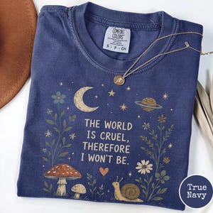 May include: A navy blue crew neck t-shirt with a graphic print featuring a crescent moon, a planet, flowers, mushrooms, and a snail. The text reads, THE WORLD IS CRUEL, THEREFORE I WON'T BE. A gold-toned necklace with a small circular pendant rests on the shirt.