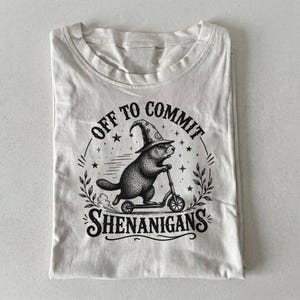 May include: A cream-colored t-shirt featuring a black graphic print of a groundhog wearing a wizard hat and riding a kick scooter. The text "OFF TO COMMIT SHENANIGANS" is arched around the illustration, which is accented with stars and leaves.