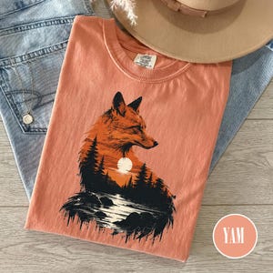 Peut inclure: Un t-shirt couleur corail avec un motif représentant la silhouette d'un renard remplie d'une scène de forêt, de lac et de coucher de soleil. Le t-shirt possède un col rond et une étiquette Comfort Colors. Le design montre un renard orange.