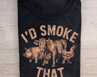 May include: A black crew neck t-shirt featuring a graphic illustration of a pig, a cow, and a chicken. The text "I'D SMOKE THAT" is printed in a distressed, tan-colored font arched above and below the animals on the front of the garment.
