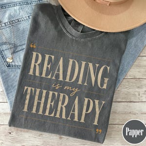 Puede incluir: Una camiseta de cuello redondo gris carbón con el texto "READING is my THERAPY" en una fuente beige desgastada, enmarcada por comillas y líneas horizontales. La camiseta se muestra junto a una prenda de mezclilla azul claro y un sombrero beige.